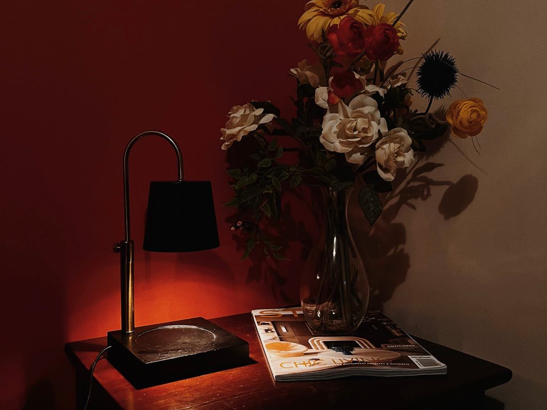 candle warmer lamp in a bedroom setting on top of a bedside table. it’s next to a flower bouquet in a warm ambient light