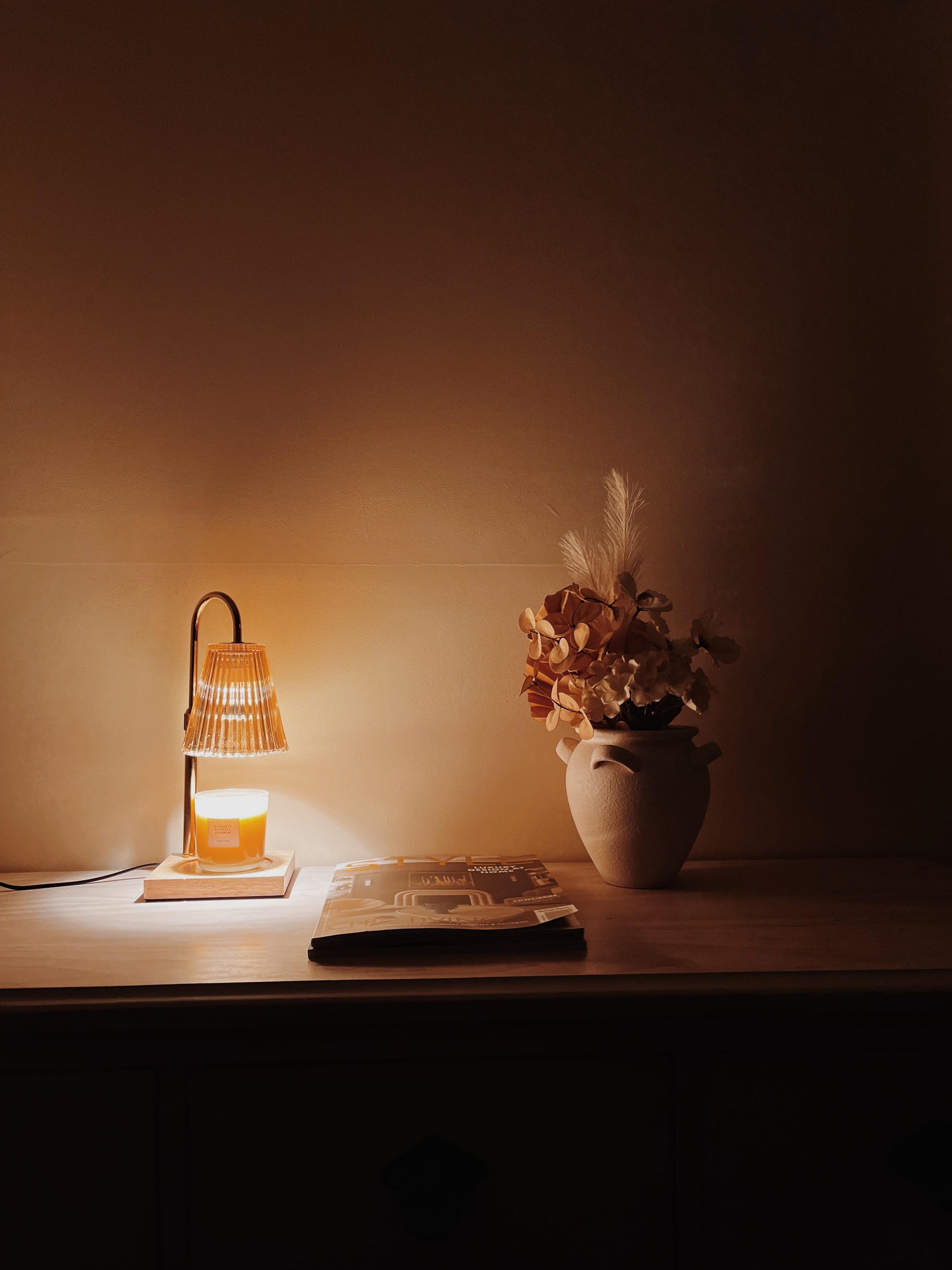 Auren Elegance Candle Warmer softly illuminating a cozy wooden tabletop with an autumnal dried flower arrangement and a design magazine, creating a serene home ambiance.