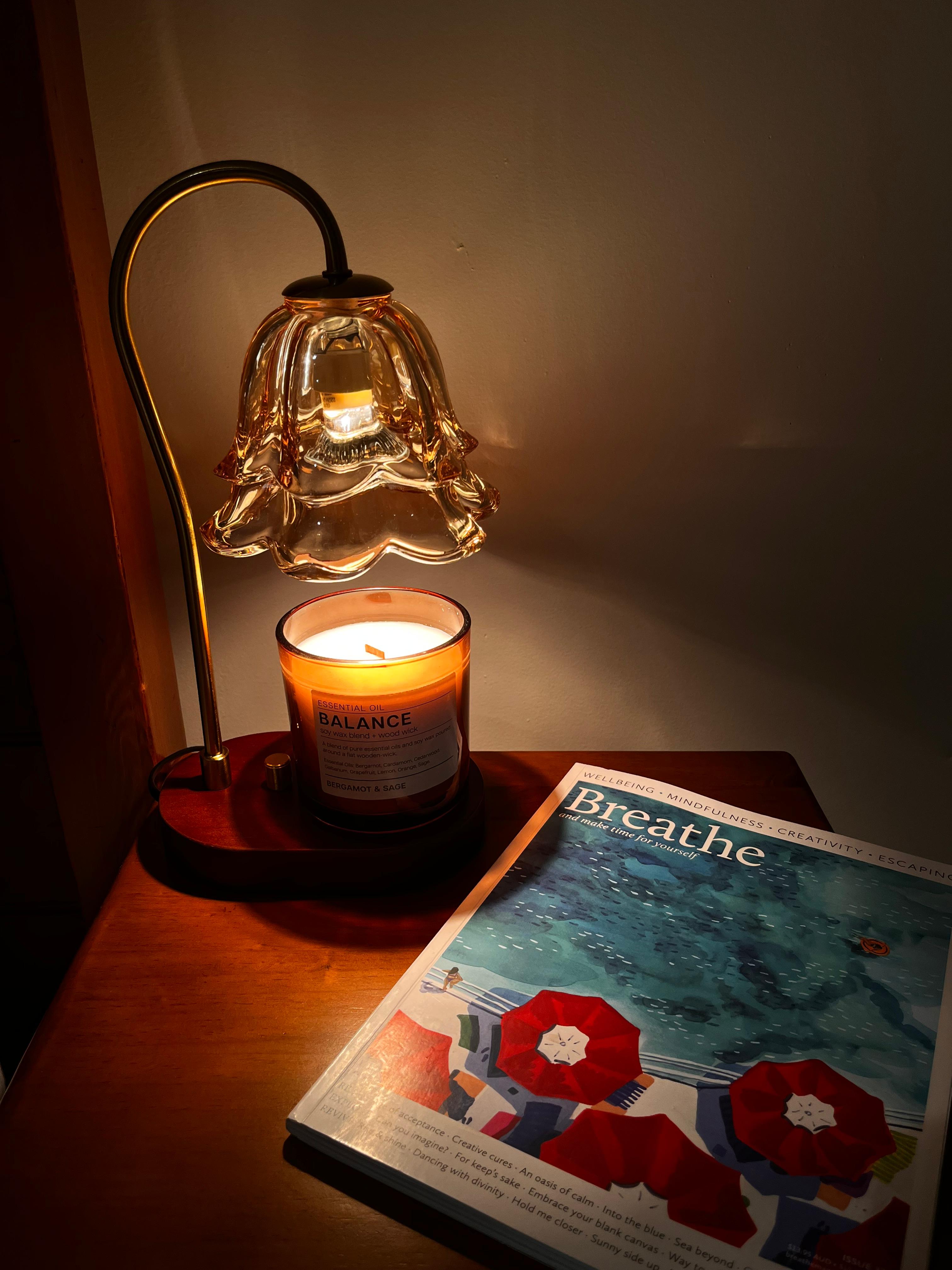 Auren Bloom candle warmer with a glowing bulb gently heats a Balance essential oil candle on a wooden nightstand, accompanied by a “Breathe” magazine—creating a serene, calming setup perfect for mindfulness and relaxation.