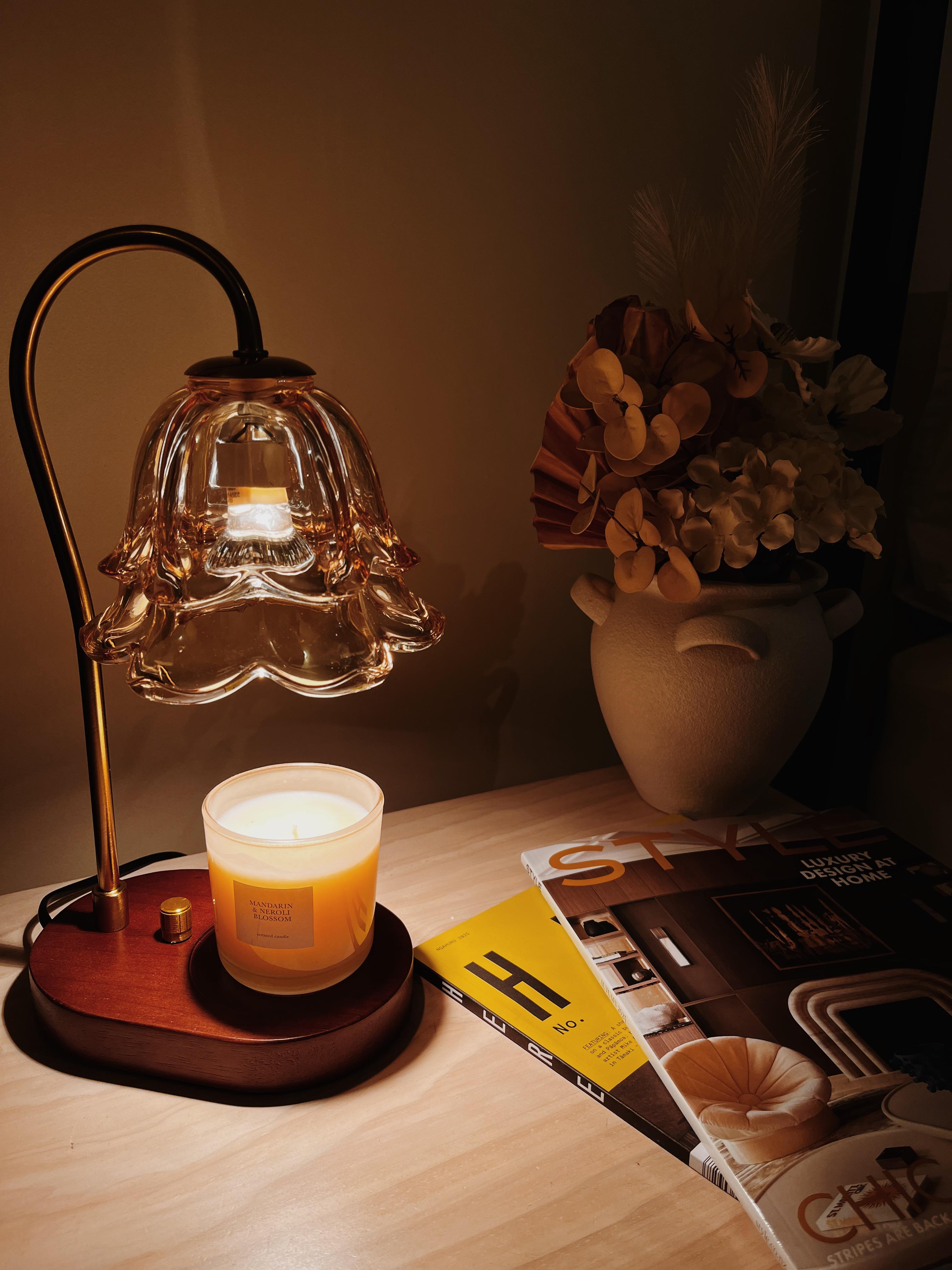 Auren Bloom candle warmer lamp with a flower-shaped glass shade warming a Mandarin Blossom candle beside style magazines and a neutral-toned dried flower arrangement, perfect for a cozy reading corner or bedside table