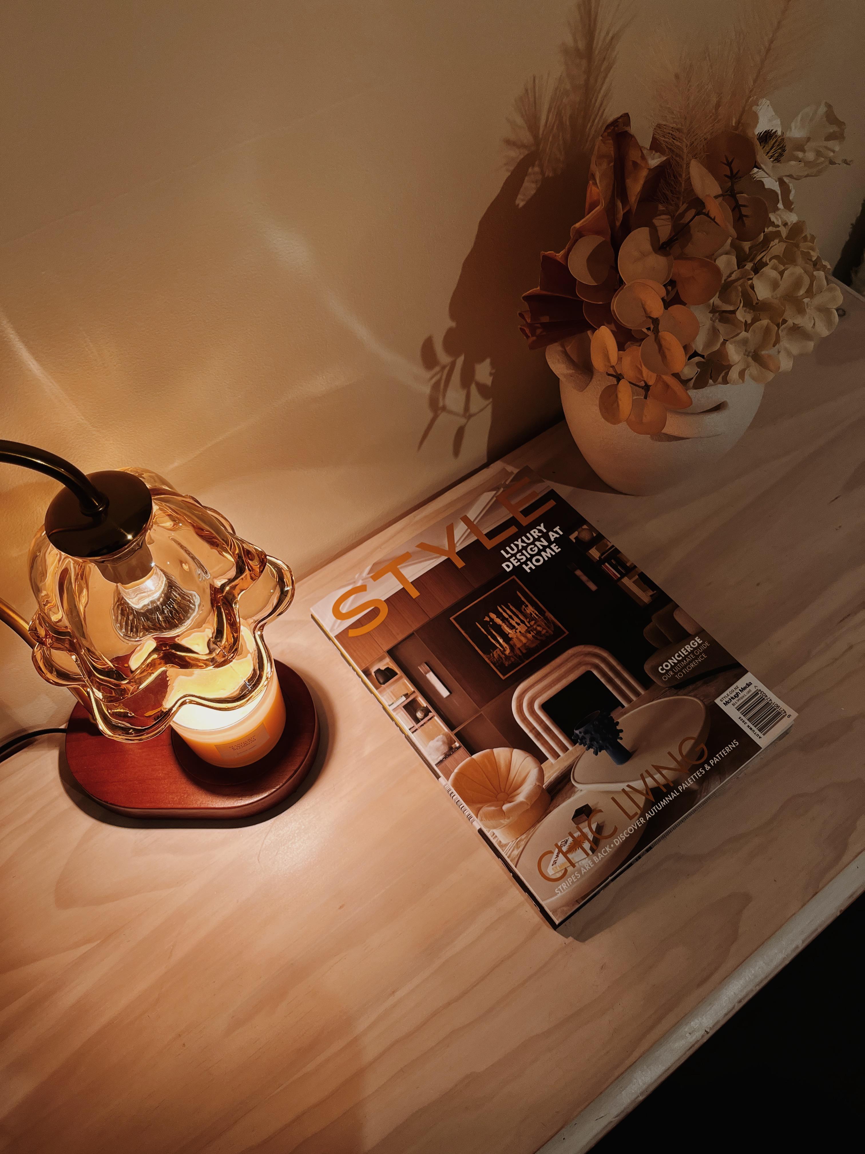 Auren Bloom Candle warmer lamp glowing softly beside a chic home magazine and a vase of dried autumn flowers, creating a warm and stylish nightstand or reading nook atmosphere
