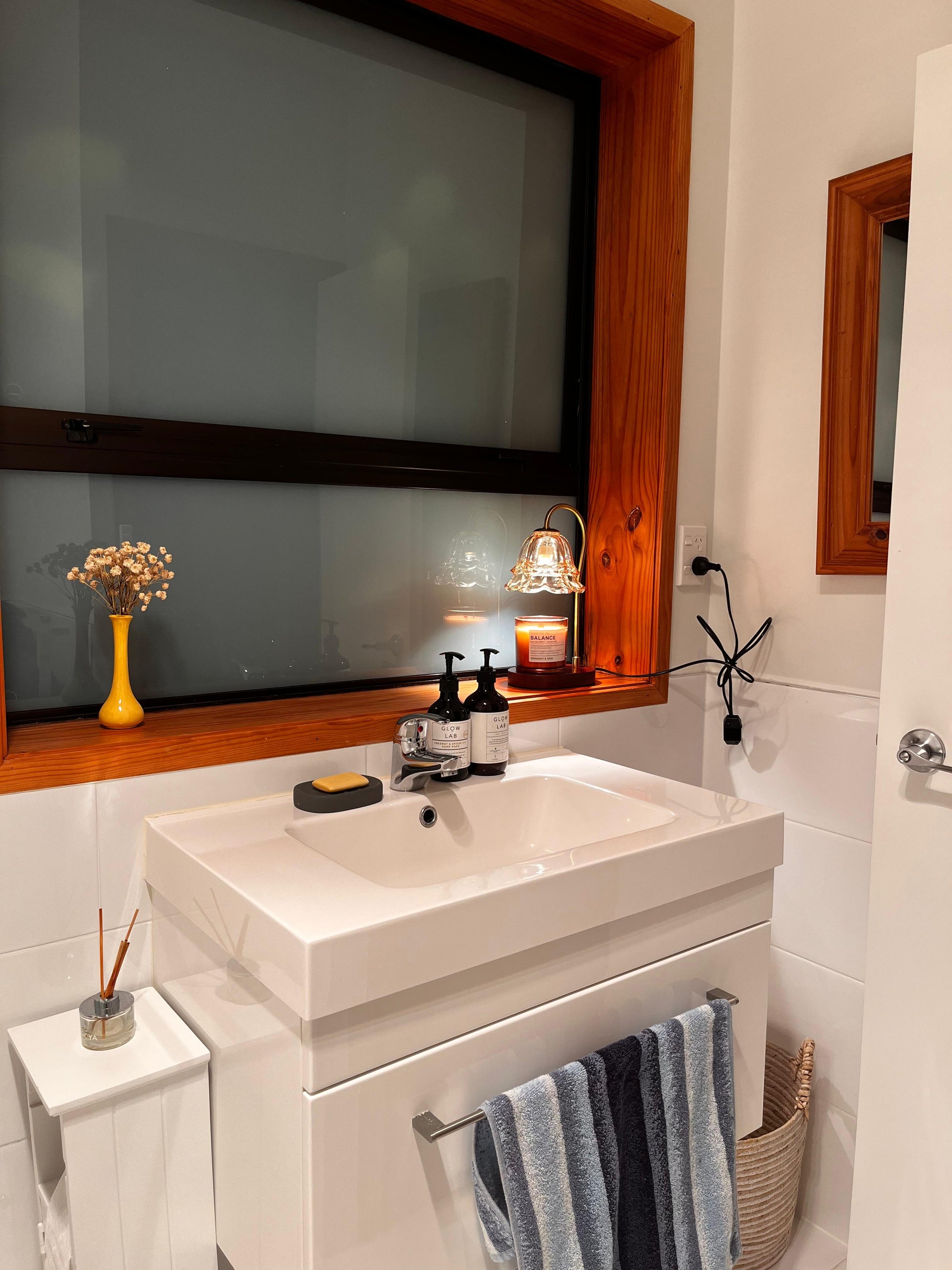 Auren Bloom Candle Warmer within a sleek, minimalist bathroom styled with soft ambient lighting from a candle warmer placed on the windowsill, illuminating a soothing scented candle and elevating the evening self-care routine with a warm, peaceful glow.