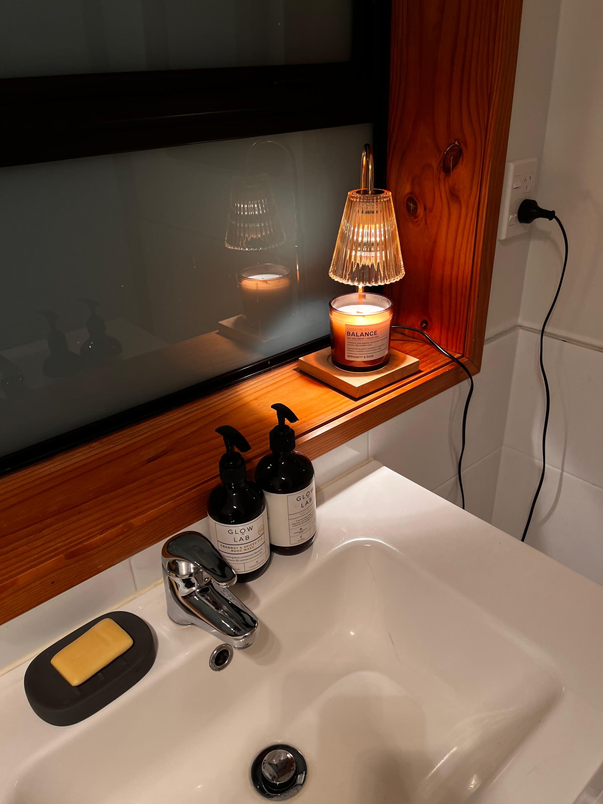 Auren Elegance Candle Warmer in a cozy bathroom scene featuring a glowing candle warmer gently melting a scented candle on the windowsill, casting a warm light over a modern white sink and natural wood accents—ideal for creating a tranquil, spa-like retreat at home.
