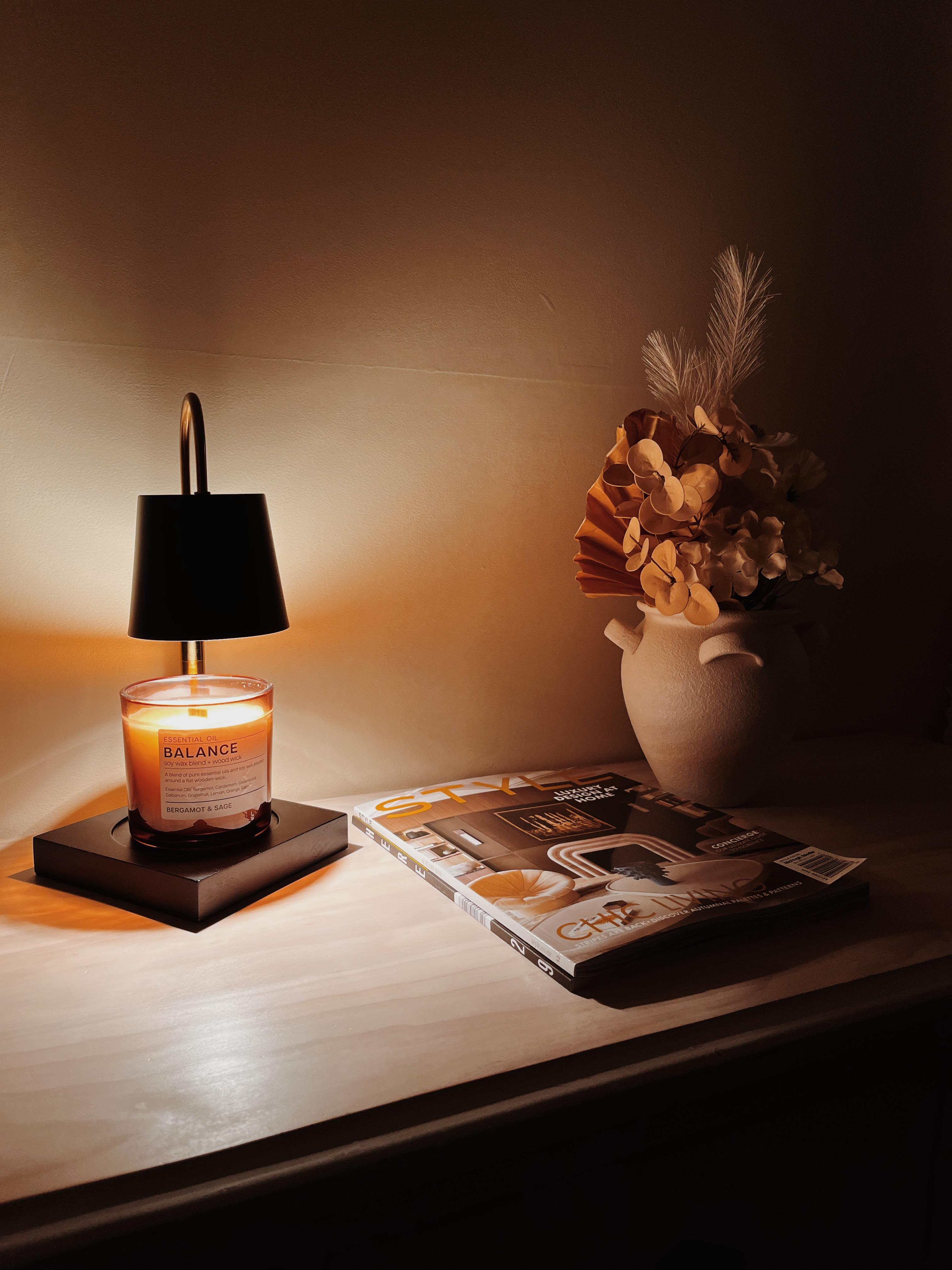 Auren Modern black candle warmer melting a Balance essential oil candle with bergamot and sage, placed on a light wooden surface. Styled alongside a neutral ceramic vase with dried pampas and eucalyptus, and a ‘Style’ interior magazine featuring chic home decor. The soft, ambient glow enhances the serene, minimalist aesthetic—perfect for wellness routines and cozy interior styling.