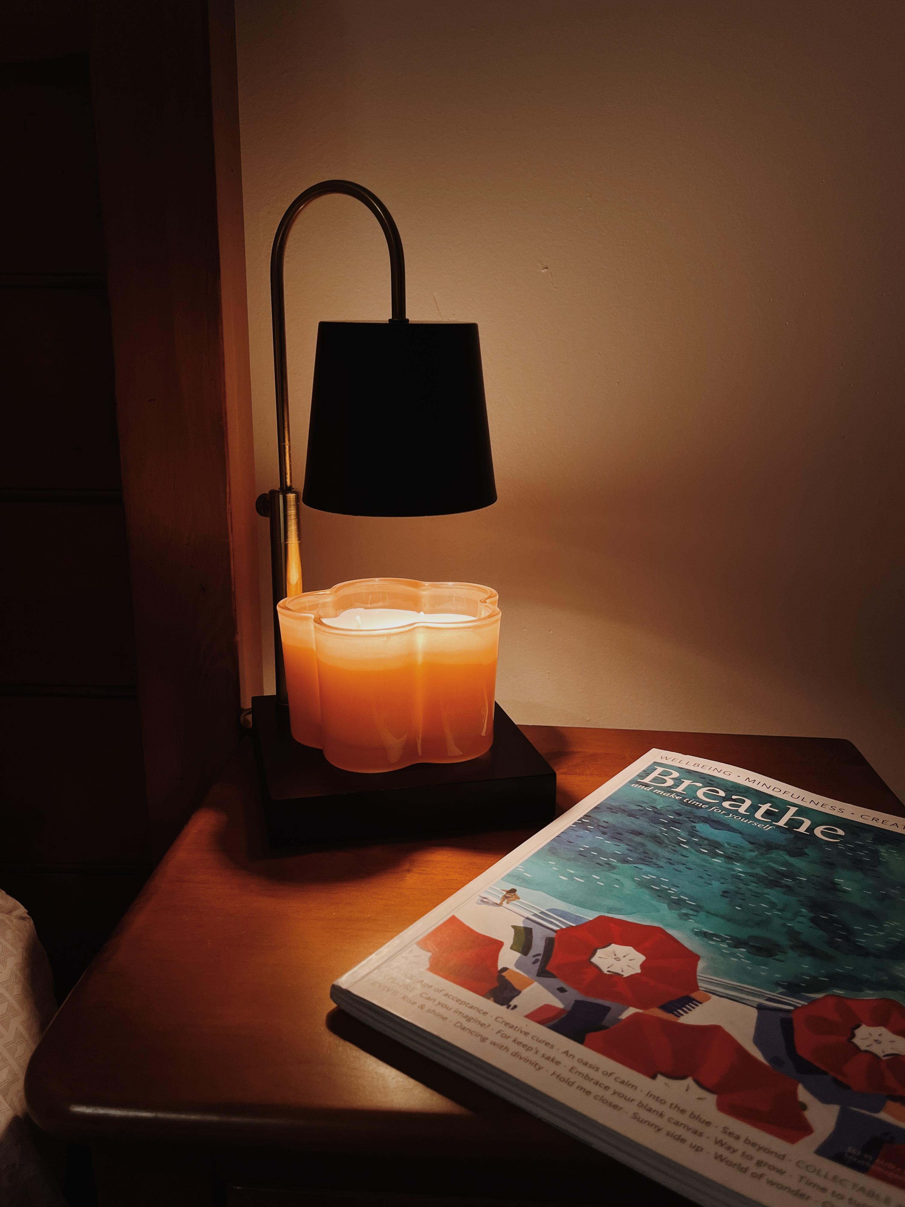 Auren Modern candle warmer melting a peach-colored flower-shaped candle on a wooden nightstand beside a Breathe magazine, creating a cozy and serene ambiance.