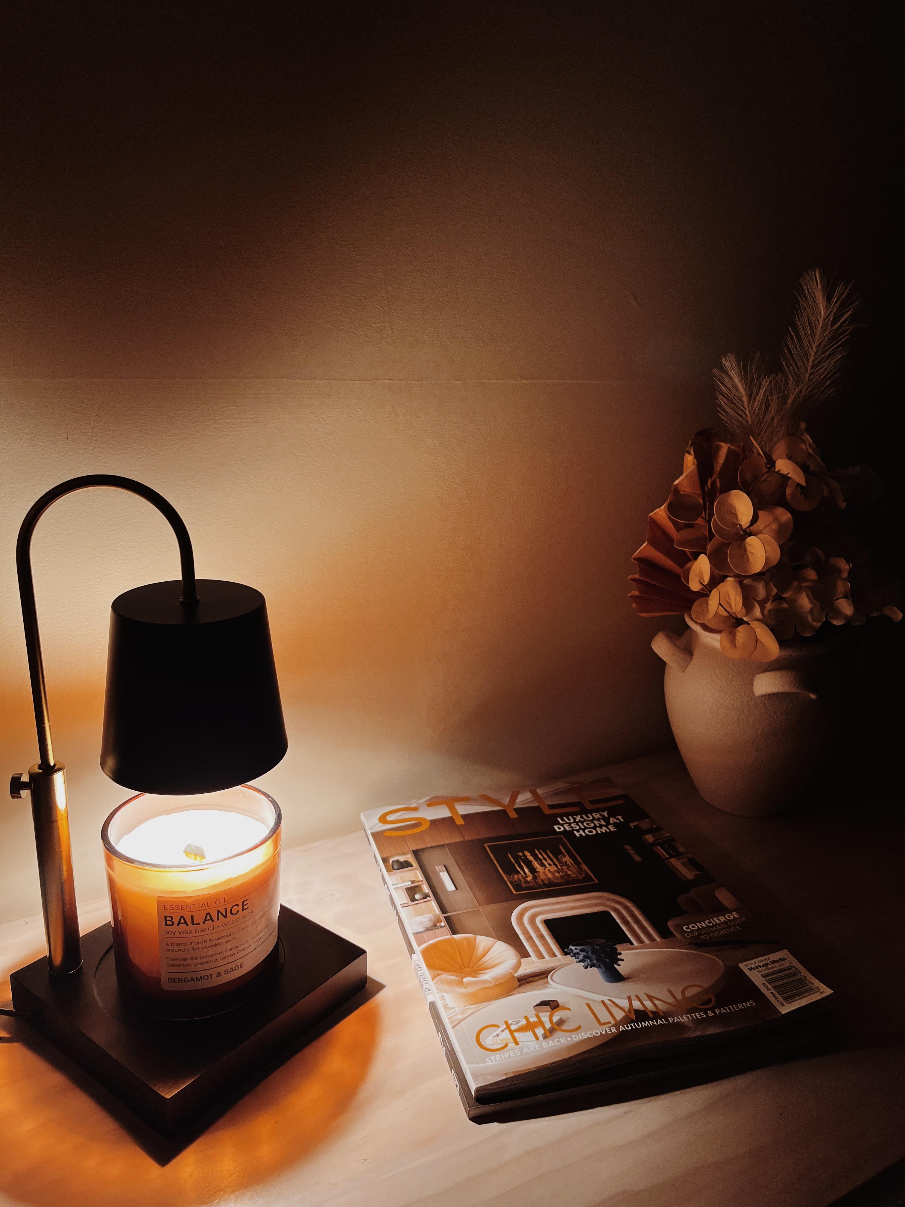 Auren Modern candle warmer gently melting a Balance essential oil candle with notes of bergamot and sage, placed on a light wooden surface beside a “Style” interior design magazine and a neutral-toned ceramic vase with dried florals. The warm ambient glow creates a calming, spa-like atmosphere perfect for wellness and cozy home decor styling.