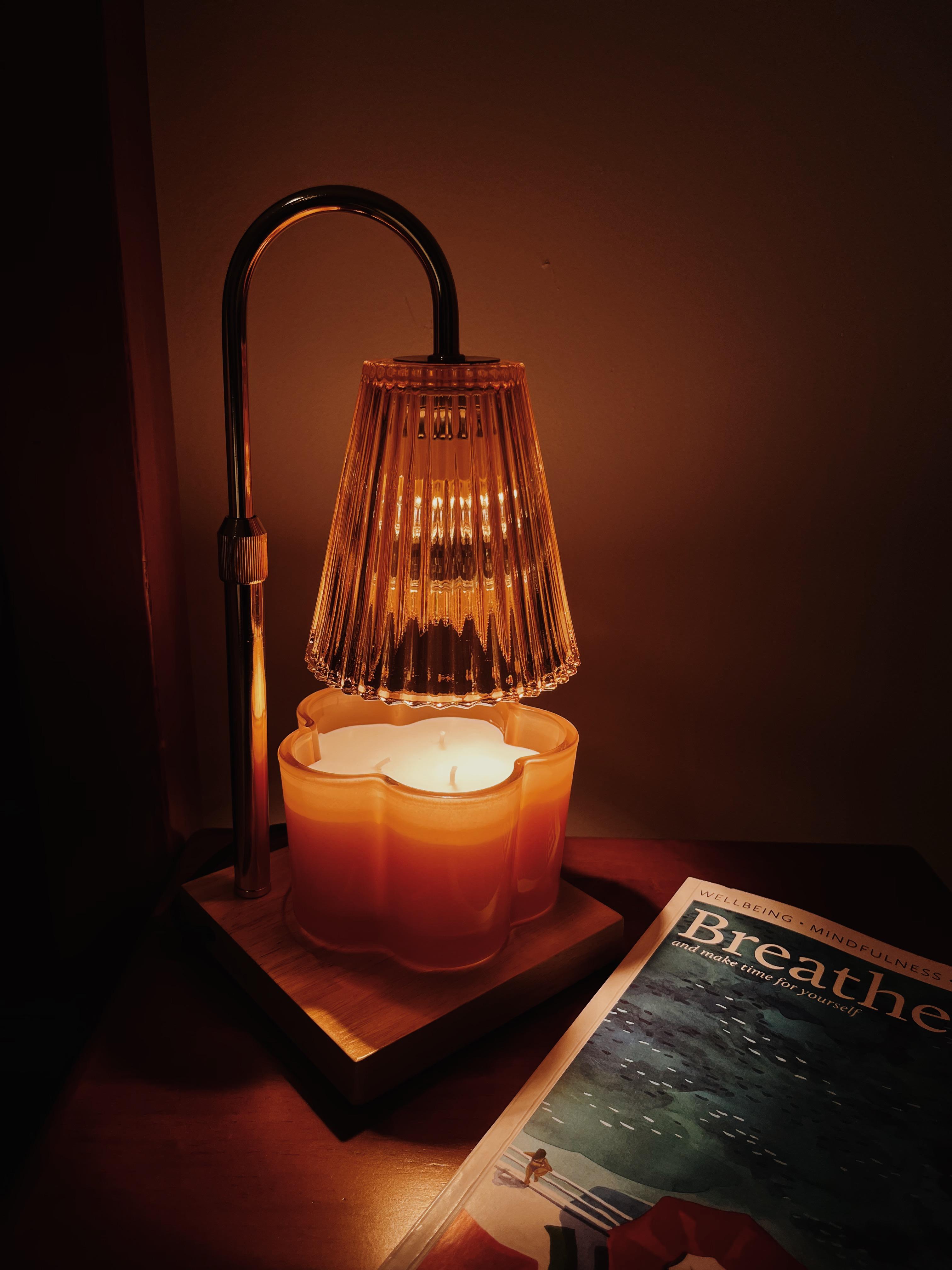 Auren Elegance Candle Warmer close up, it is glowing a warm golden orange colour as it warms a flower shaped candle. It sits on a bedside table next to a vibrant magazine. 